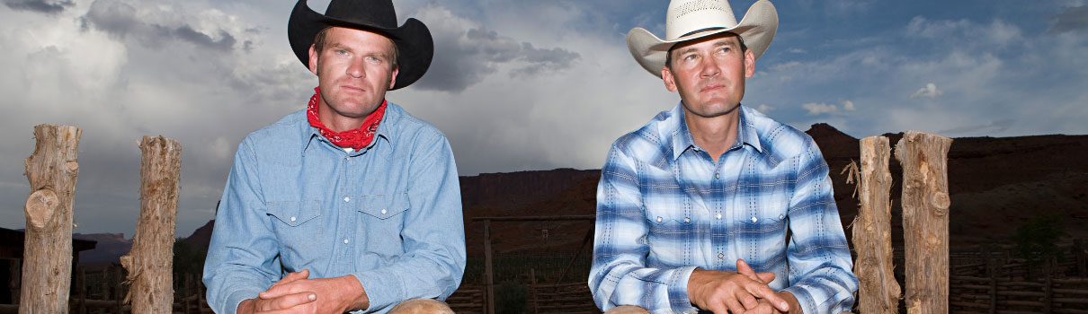Zwei Cowboys sitzen auf einem Zaun; der Linke trägt einen schwarzen, der Rechte einen weißen Hut